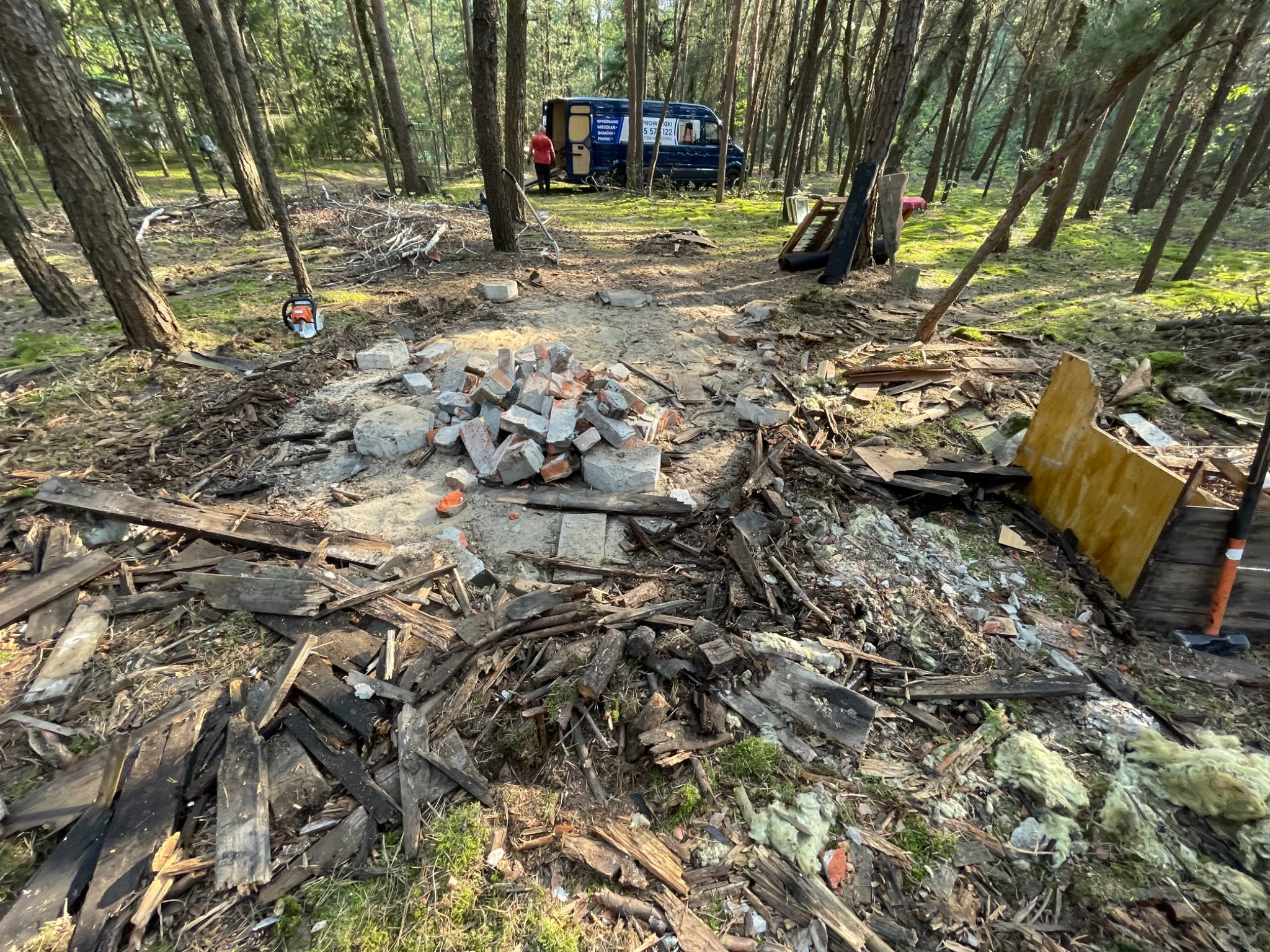 Teren po usunięciu konstrukcji w lesie, widoczne resztki drewna, cegieł, izolacji, piła łańcuchowa i samochód dostawczy w tle.