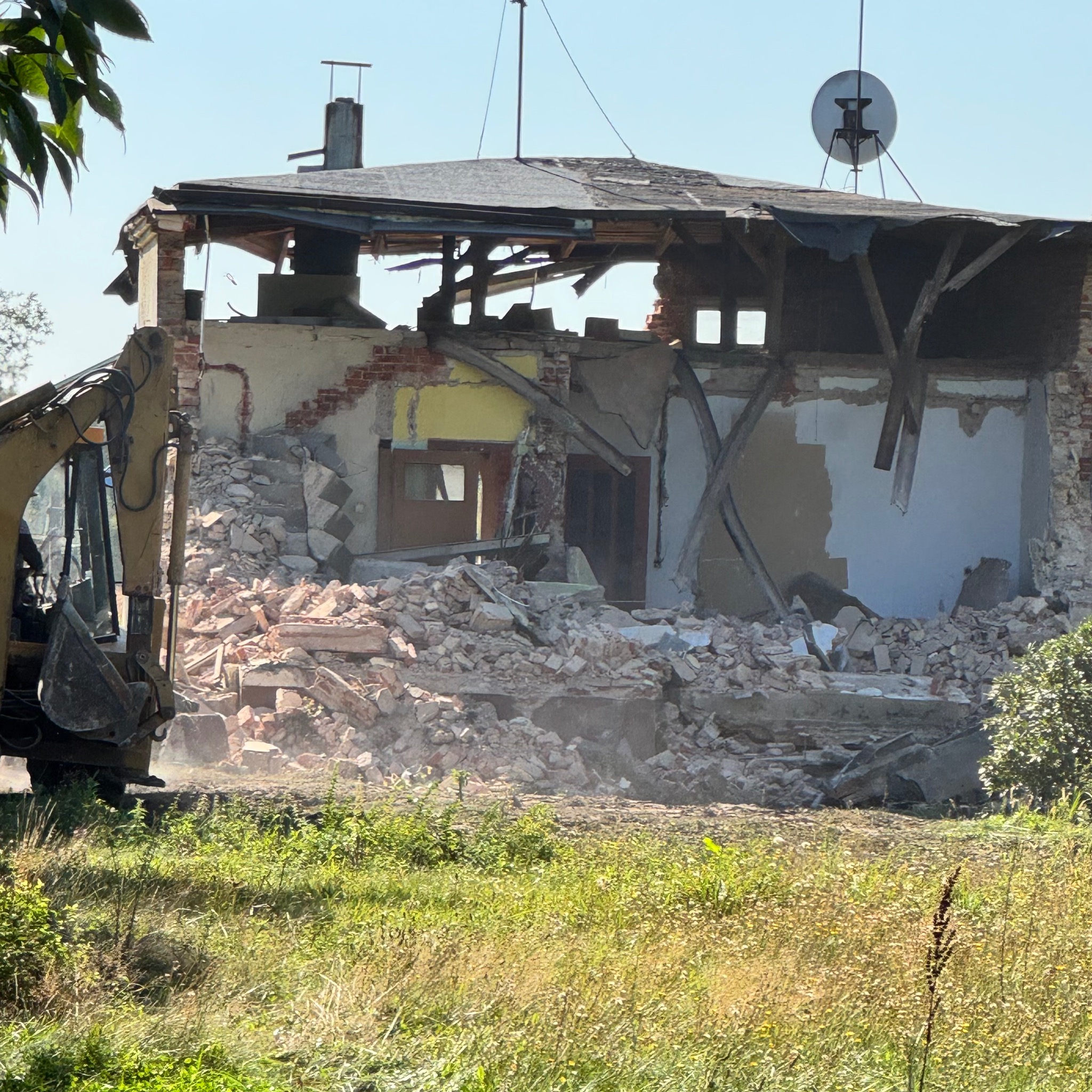 Żółta koparka częściowo widoczna z lewej strony, gruzowisko po rozbiórce budynku z widocznymi fragmentami ścian, okna i dachu, na dachu antena satelitarna, a w tle zielona łąka.