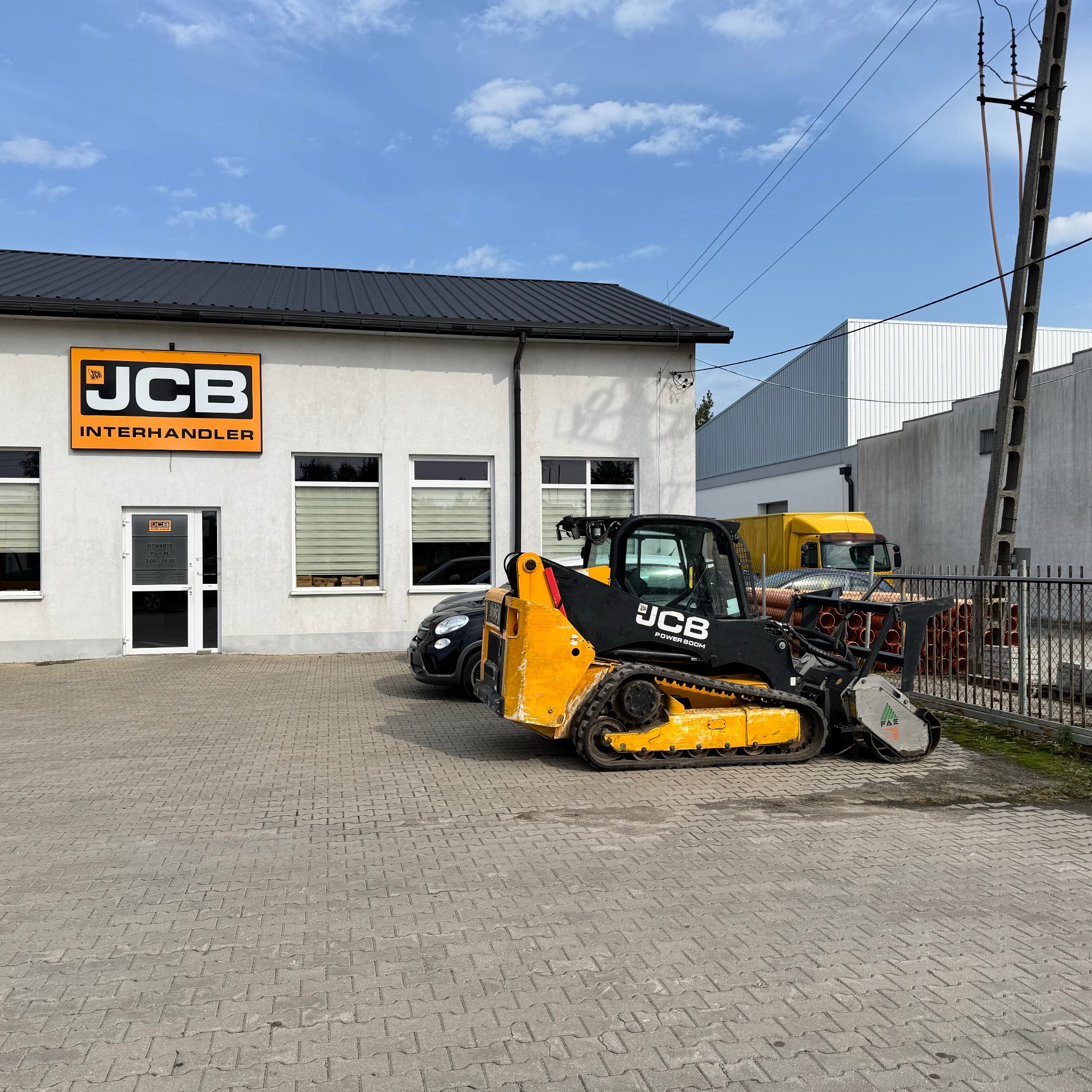 Żółto-czarna minikoparka gąsienicowa JCB z frezem do pni stoi na kostce brukowej przed budynkiem firmy JCB Interhandler w słoneczny dzień.