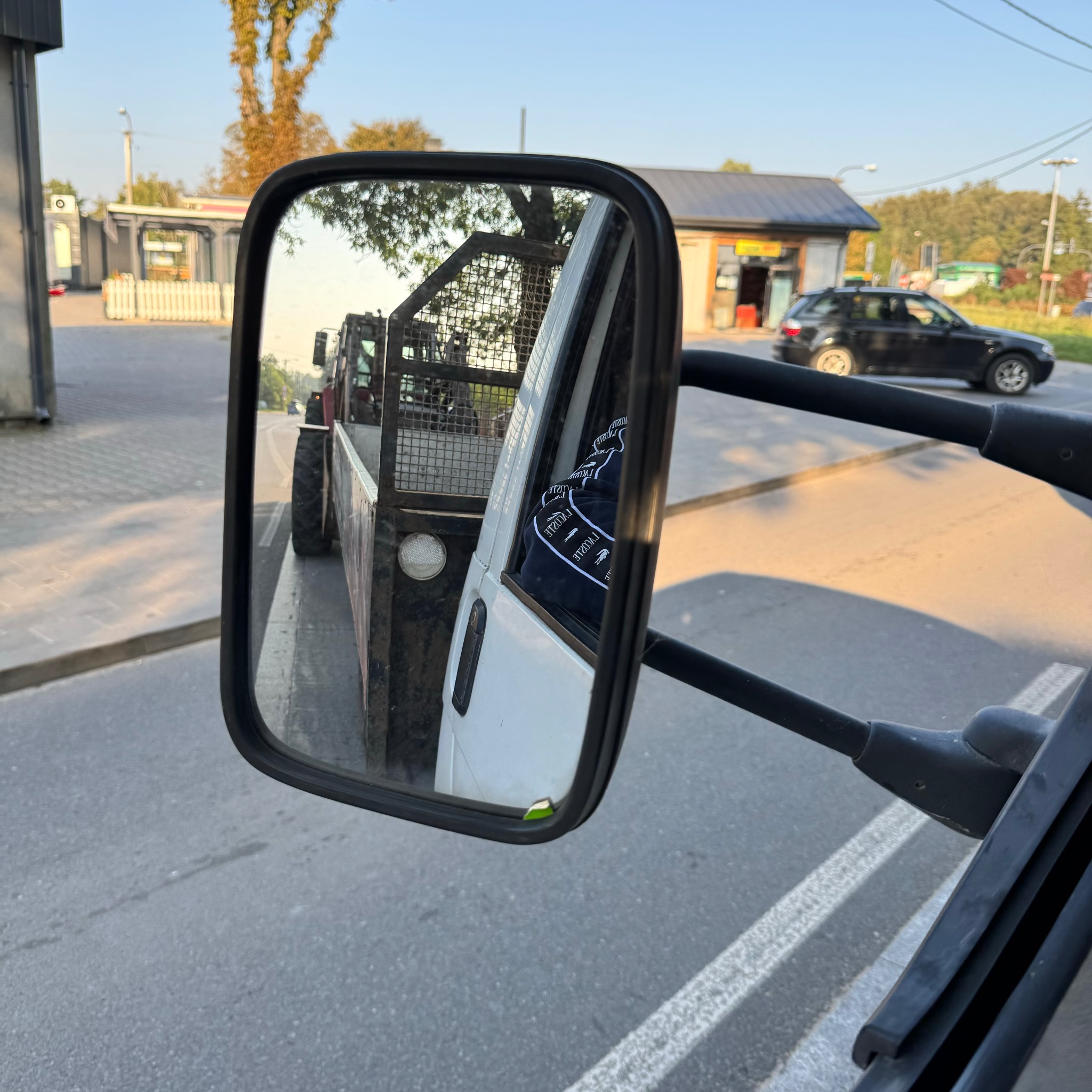 Widok w lusterku bocznym samochodu na przyczepę ciągniętą przez traktor, prawdopodobnie po wykonanej usłudze, z widoczną zabudową miejską w tle.