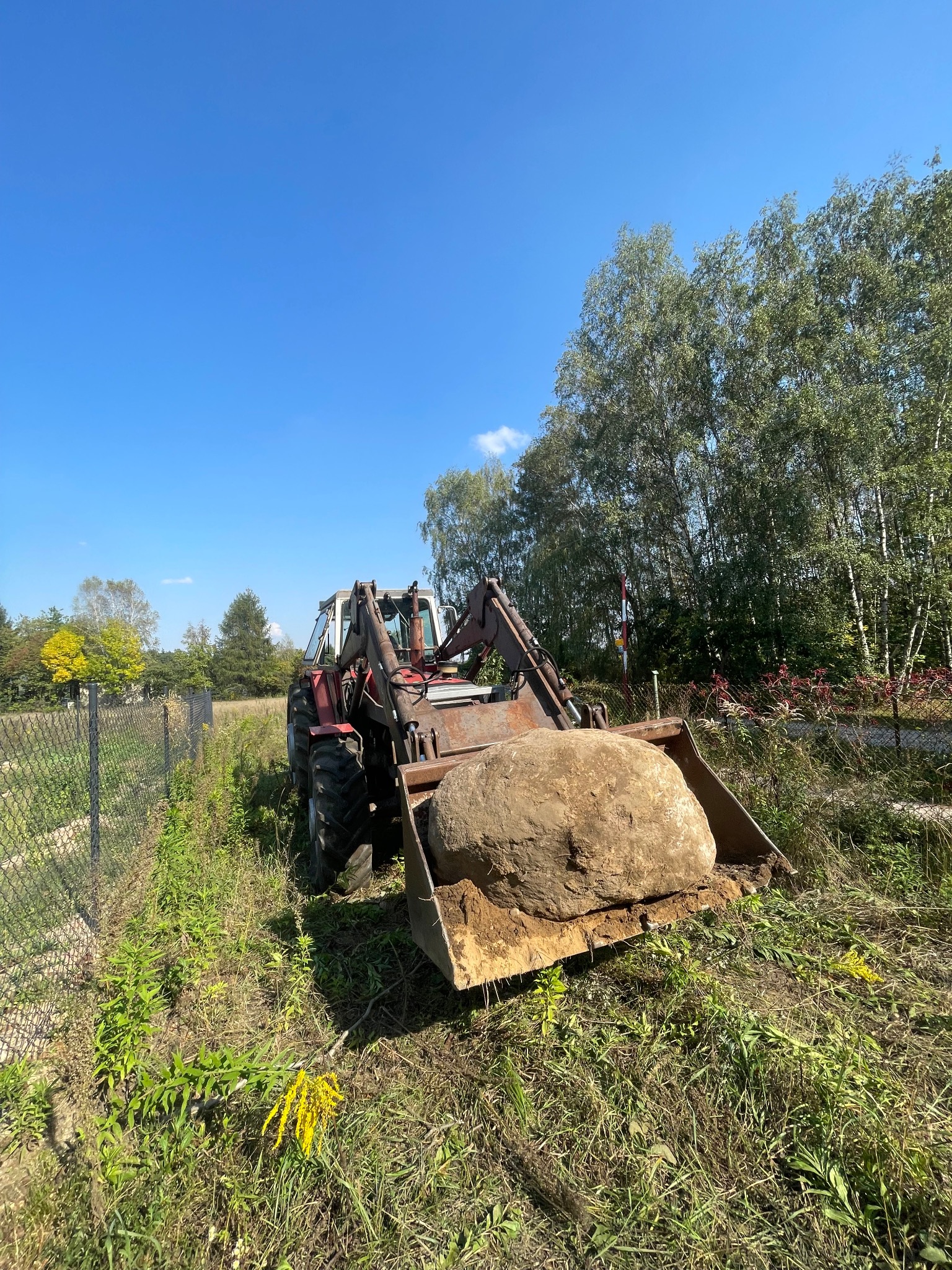 Czerwony traktor z łyżką transportuje duży, ziemisty kamień na tle zieleni i błękitnego nieba.