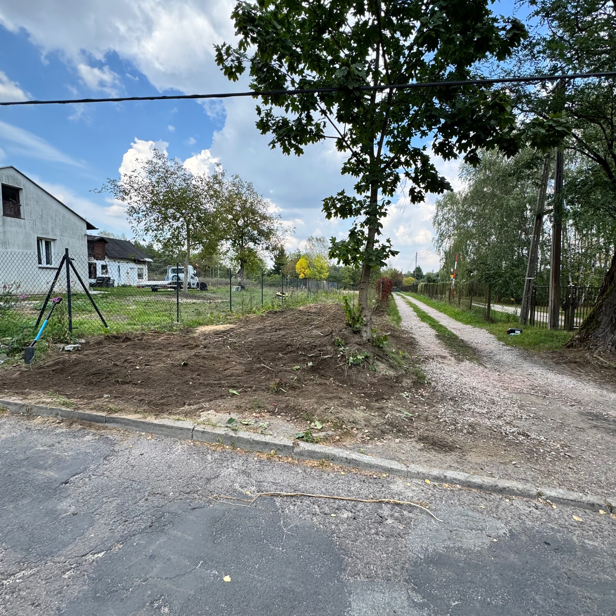 Przygotowany teren pod trawnik, świeżo przekopana ziemia, widoczne korzenie i resztki po wycince drzew, w tle budynek mieszkalny i zaparkowany samochód dostawczy.