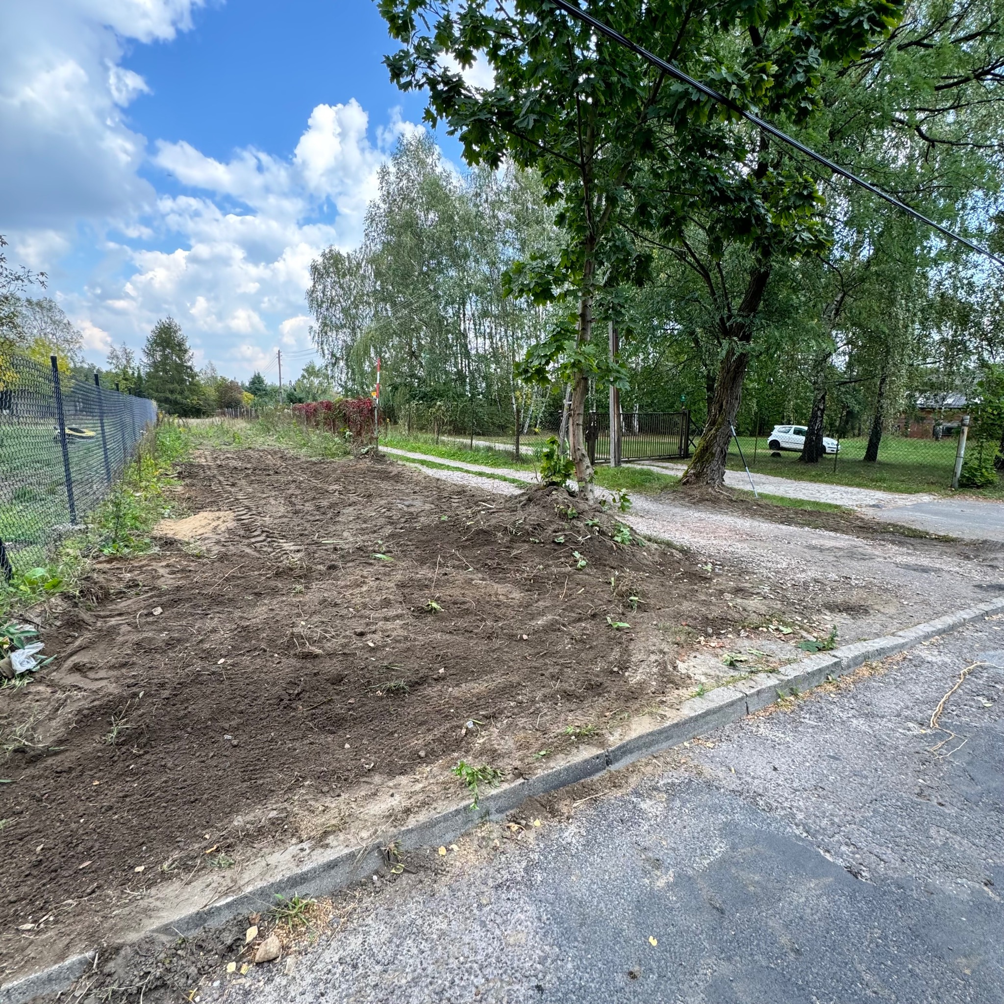 Przygotowany teren pod trawnik z widocznymi śladami opon, częściowo uprzątnięty, obok chodnik i ulica, w tle drzewa i ogrodzenie.