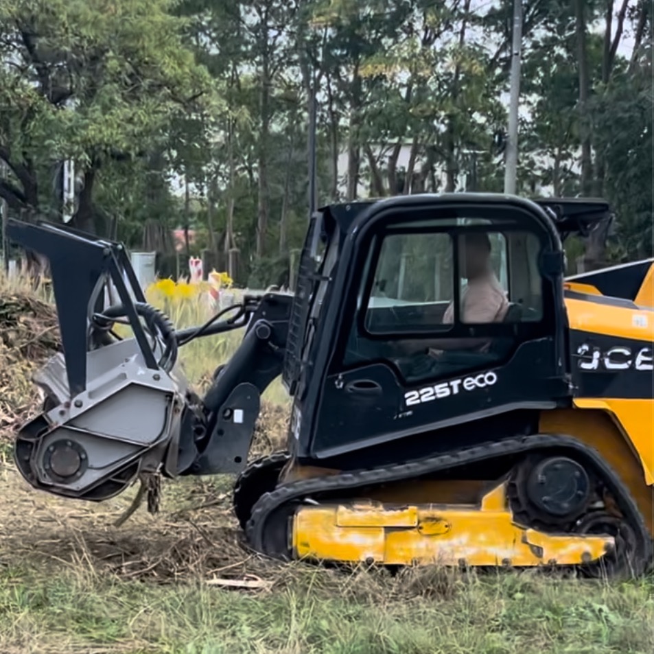 Żółto-czarna maszyna do frezowania pni drzew JCB 225T eco, z operatorem w kabinie, na zarośniętym terenie w tle drzewa.