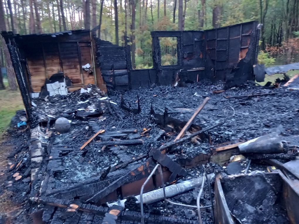 Spalony doszczętnie drewniany budynek z widocznymi resztkami ścian i fundamentów na tle lasu.