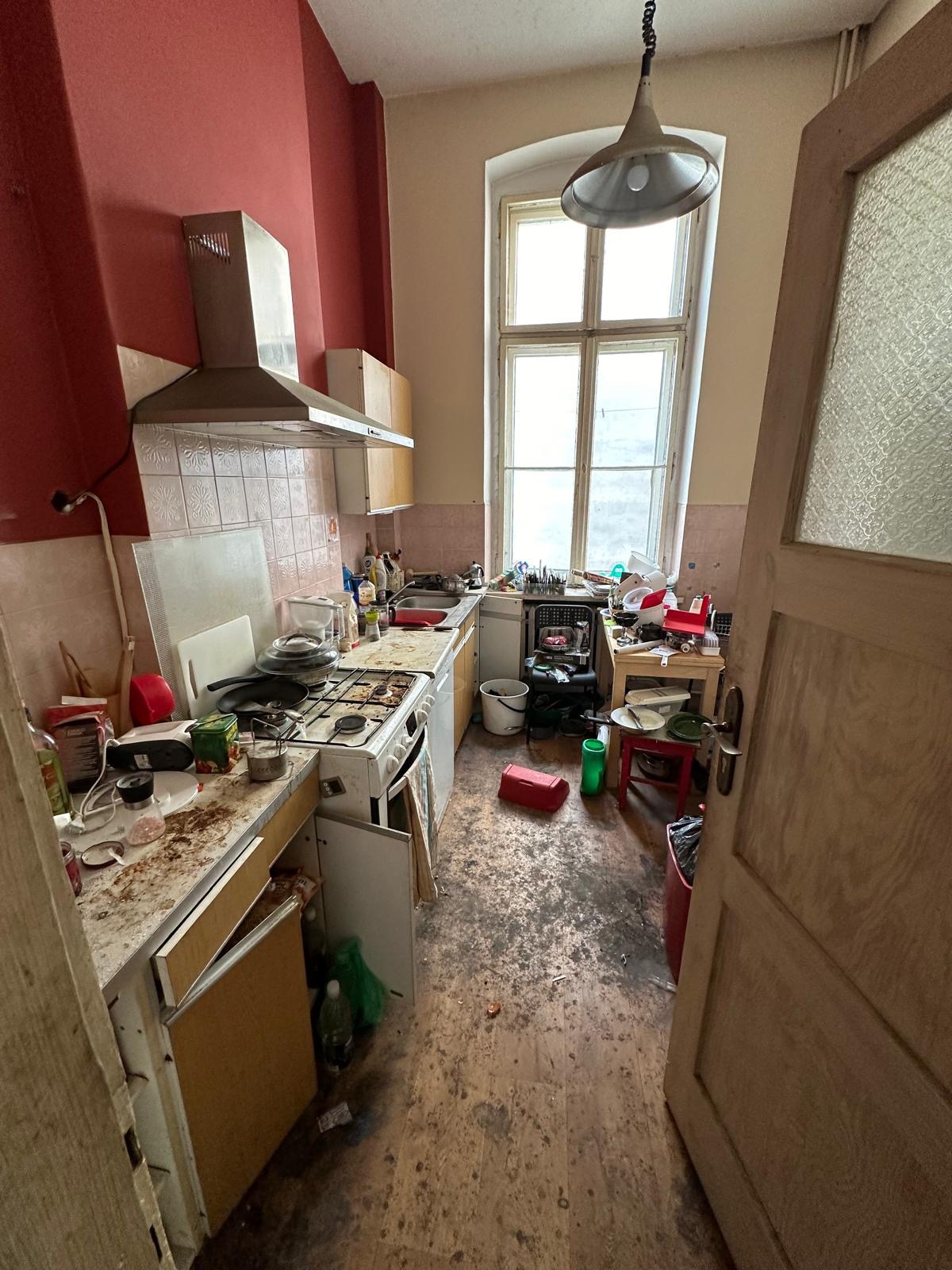 Bardzo zaniedbana kuchnia z otwartymi drzwiami, widoczne stare meble, brudne podłogi i ściany, oraz duży bałagan na blatach, stole i podłodze.