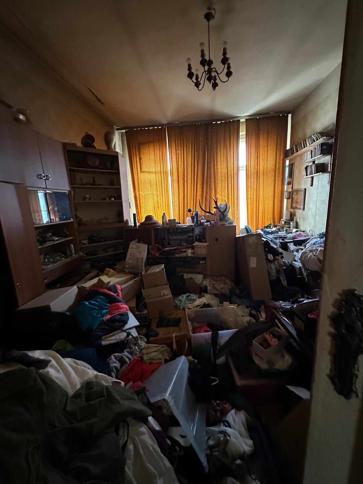 Pokój wypełniony chaotycznym nagromadzeniem przedmiotów, kartonów i ubrań, z widocznymi meblami i zasłonami w tle, oświetlony przez okno zza zasłon.