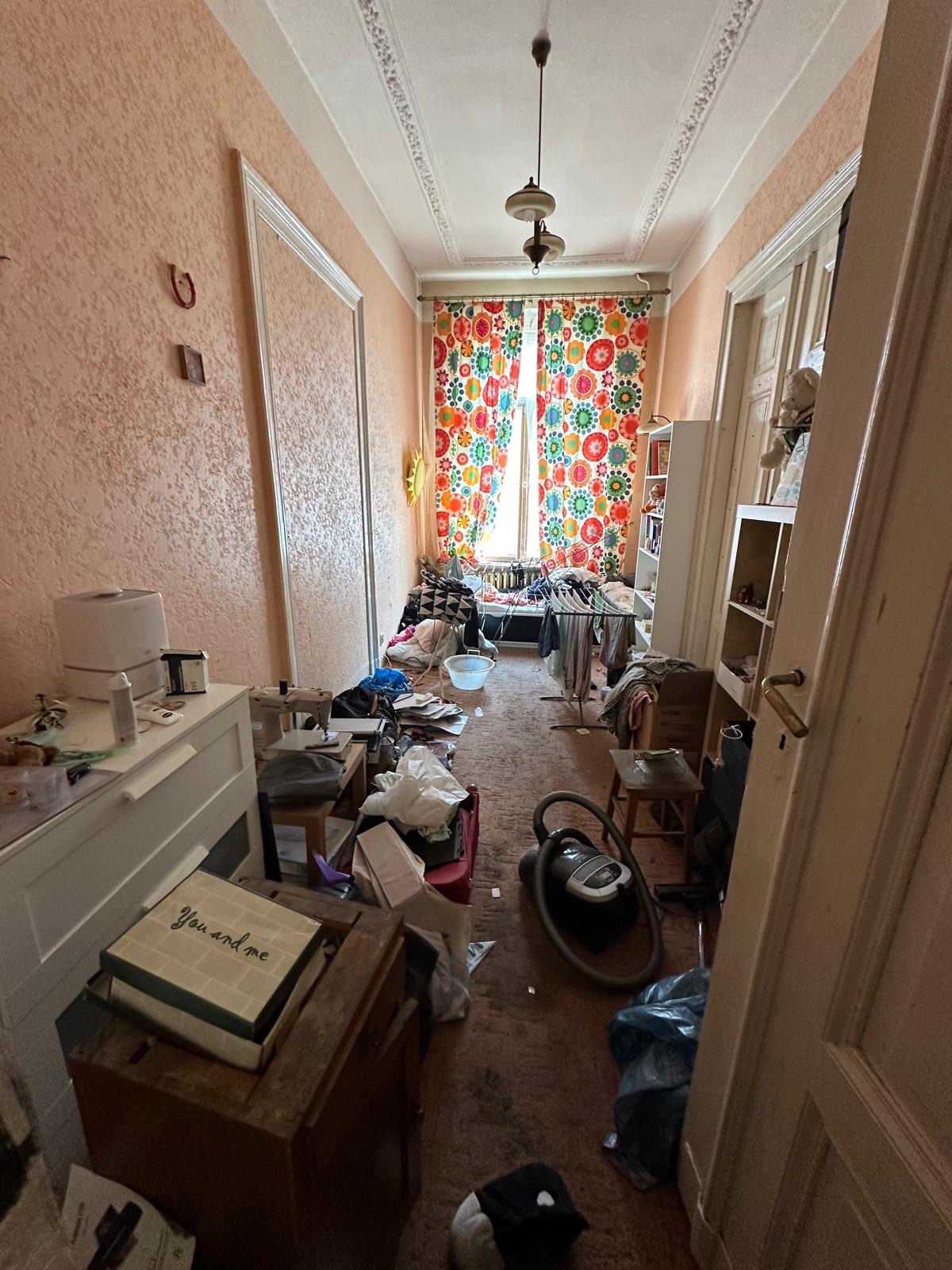 Długi, wąski korytarz zagracony meblami, kartonami, workami i sprzętami AGD, z kolorowymi zasłonami w tle, ukazujący stan pomieszczenia przed rozpoczęciem porządkowania.