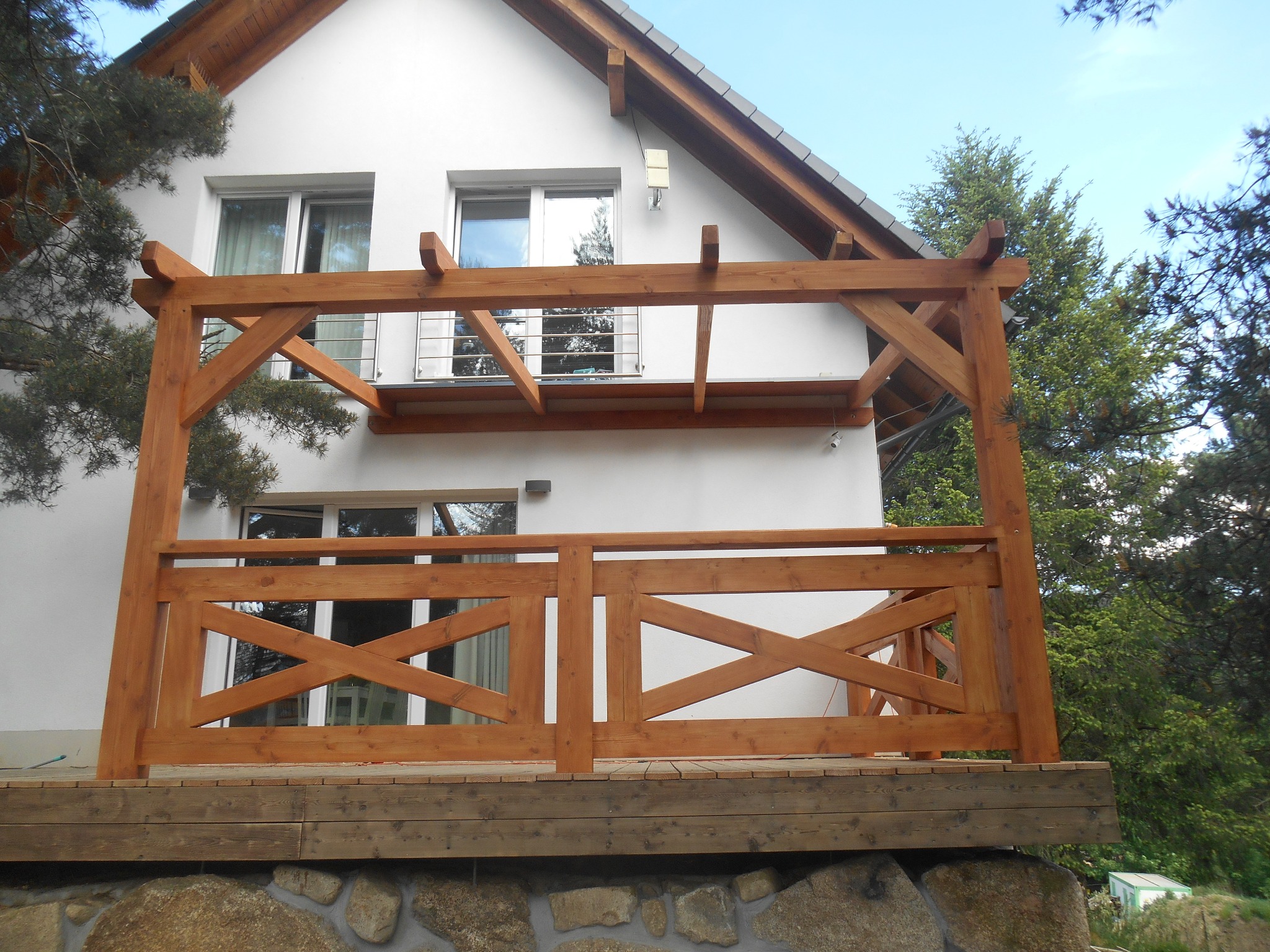 Drewniany taras z balustradą w kształcie krzyży, przylegający do białego domu z widocznymi oknami i szarą dachówką, osadzony na kamiennej podmurówce.