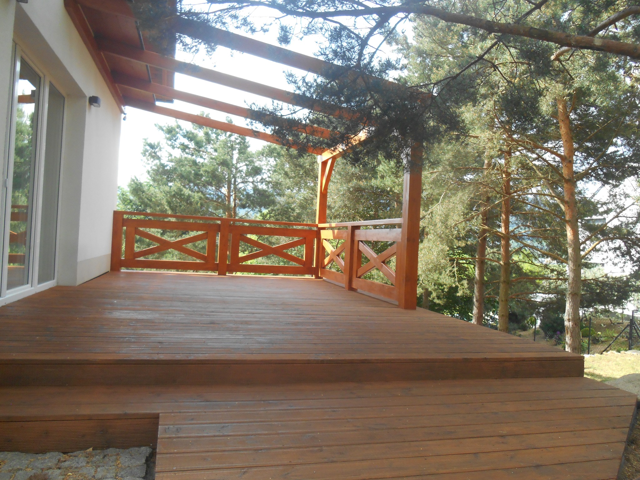 Brązowy taras z drewnianą balustradą z motywem krzyża, widok z dołu, w tle iglaki i biała ściana budynku z dużym oknem.