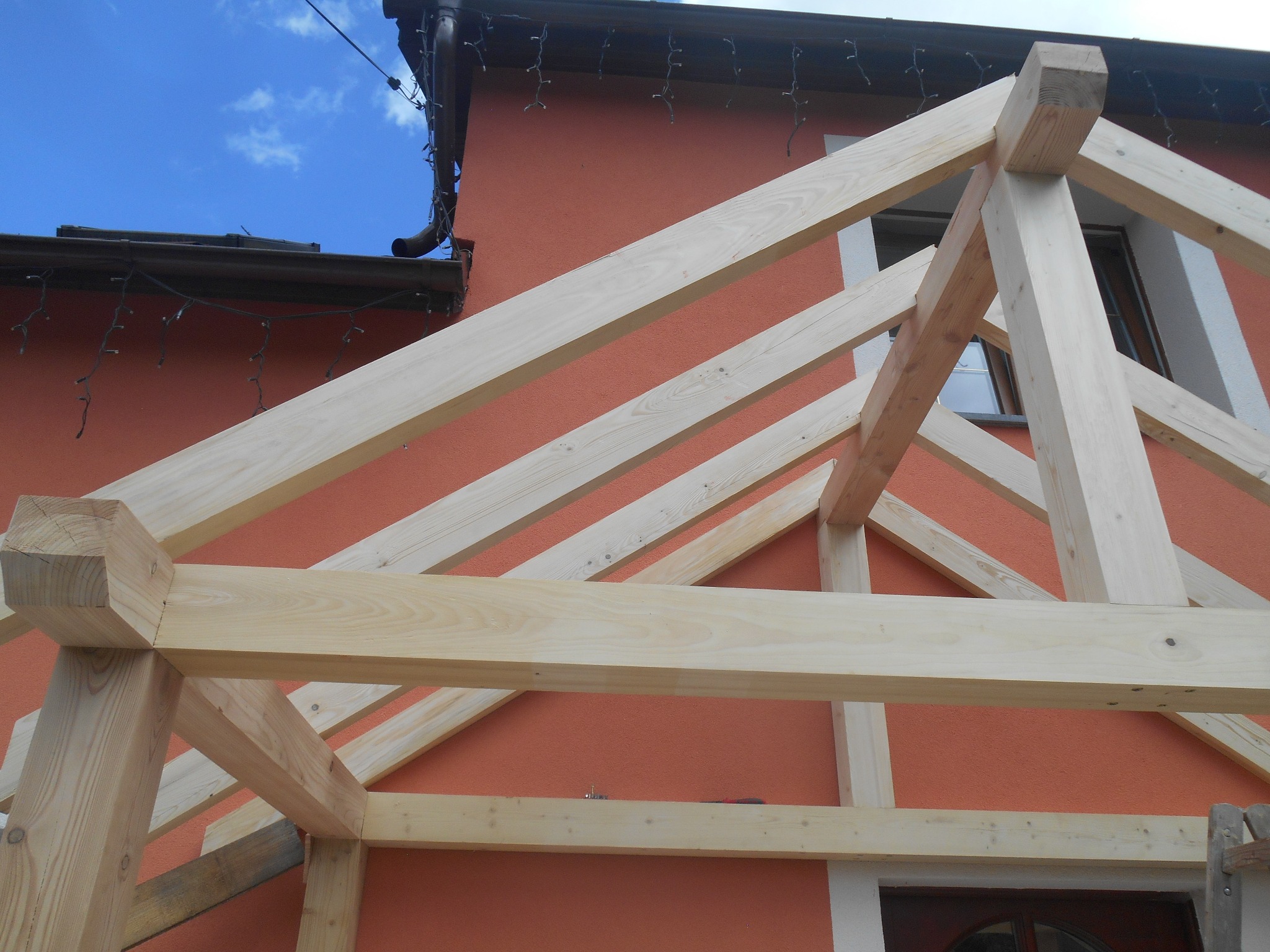 Konstrukcja drewniana z jasnych belek na tle pomarańczowej elewacji budynku, widoczne złączenia belek i fragment okna.