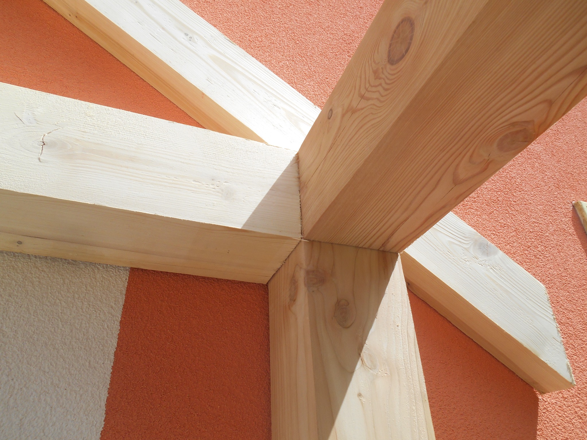 Fragment konstrukcji drewnianej daszku tarasu w trakcie budowy, widoczne połączenie belek na tle pomarańczowej elewacji.