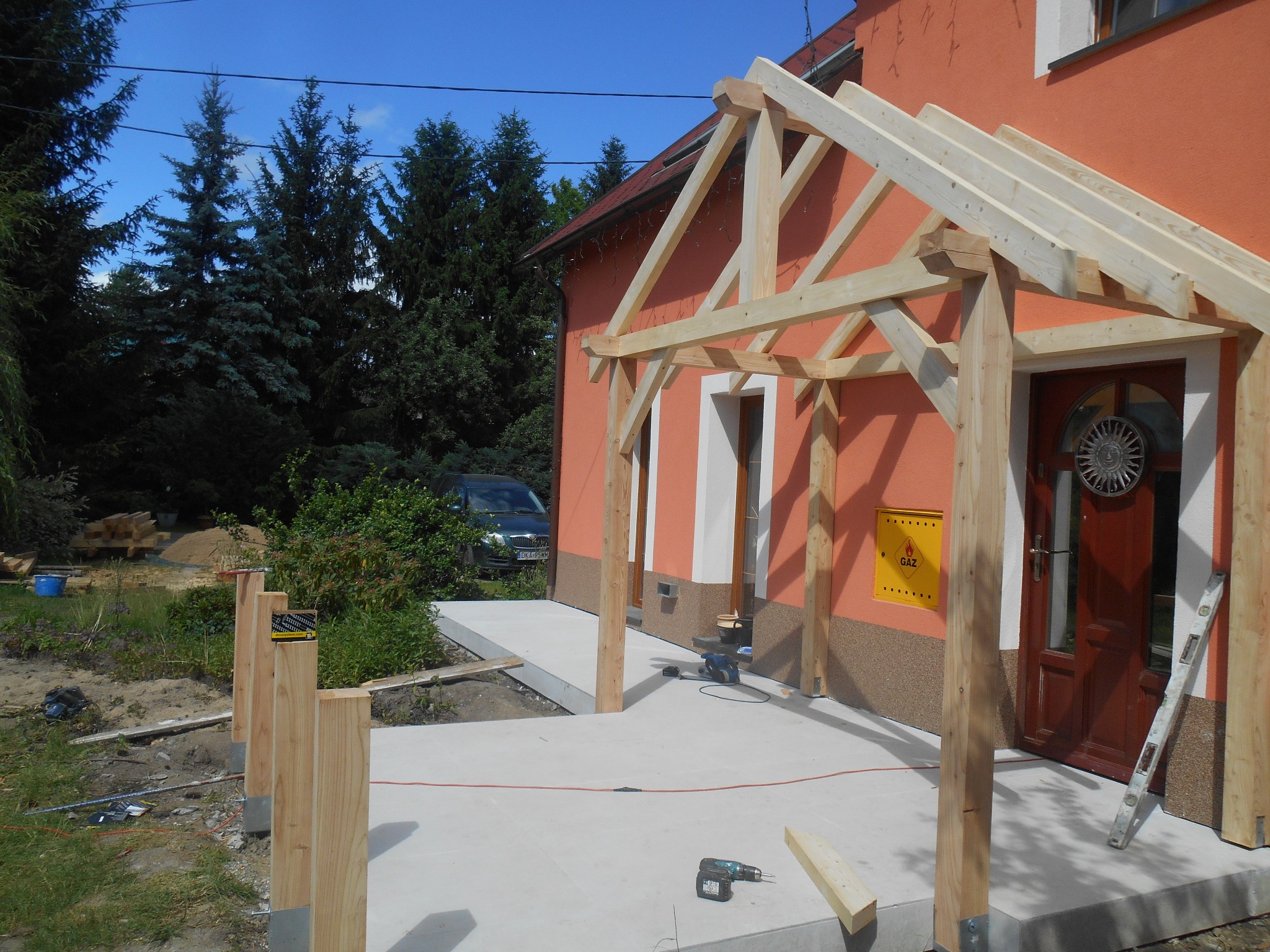 Wznoszona drewniana konstrukcja zadaszenia nad wejściem do pomarańczowego domu, widoczne narzędzia pracy, poziomica oparta o ścianę, w tle zielone drzewa i fragment samochodu.