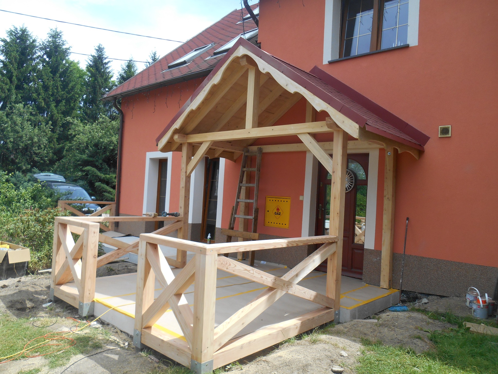 W Janowicach Wielkich trwa budowa drewnianego zadaszenia nad wejściem do domu, widoczna surowa konstrukcja z belek i desek oraz częściowo zamontowana balustrada, teren wokół w trakcie prac...