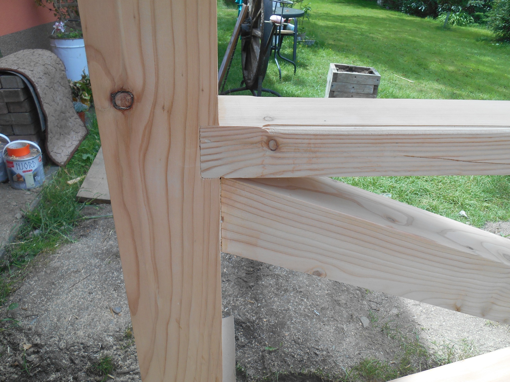 Fragment drewnianej konstrukcji w trakcie budowy, widoczne połączenia belek i słupów, w tle trawnik i elementy ogrodowe.