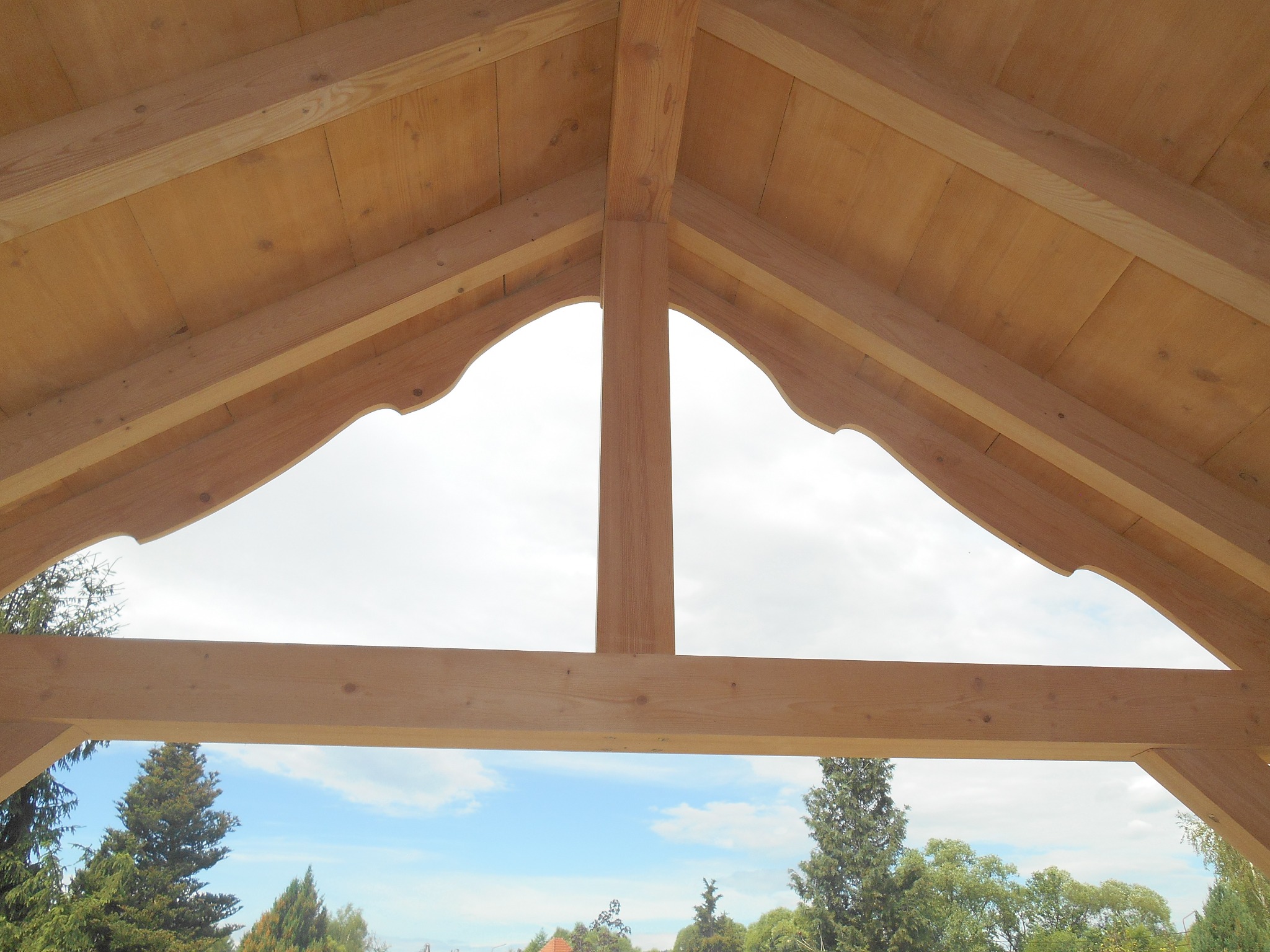 Drewniana konstrukcja dachu z widokiem na drzewa i niebo, elementy wykończone dekoracyjnymi wycięciami.