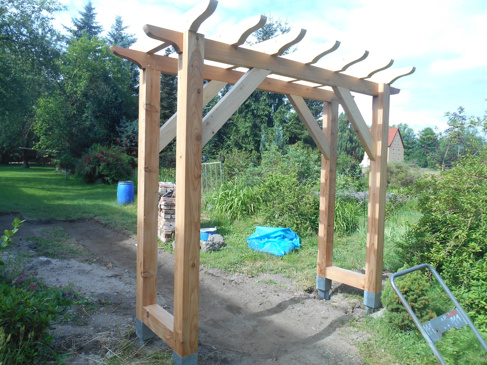 Surowa drewniana konstrukcja pergoli w ogrodzie, widoczne elementy złączone śrubami, przygotowane podłoże z wytyczoną ścieżką, narzędzia w tle.