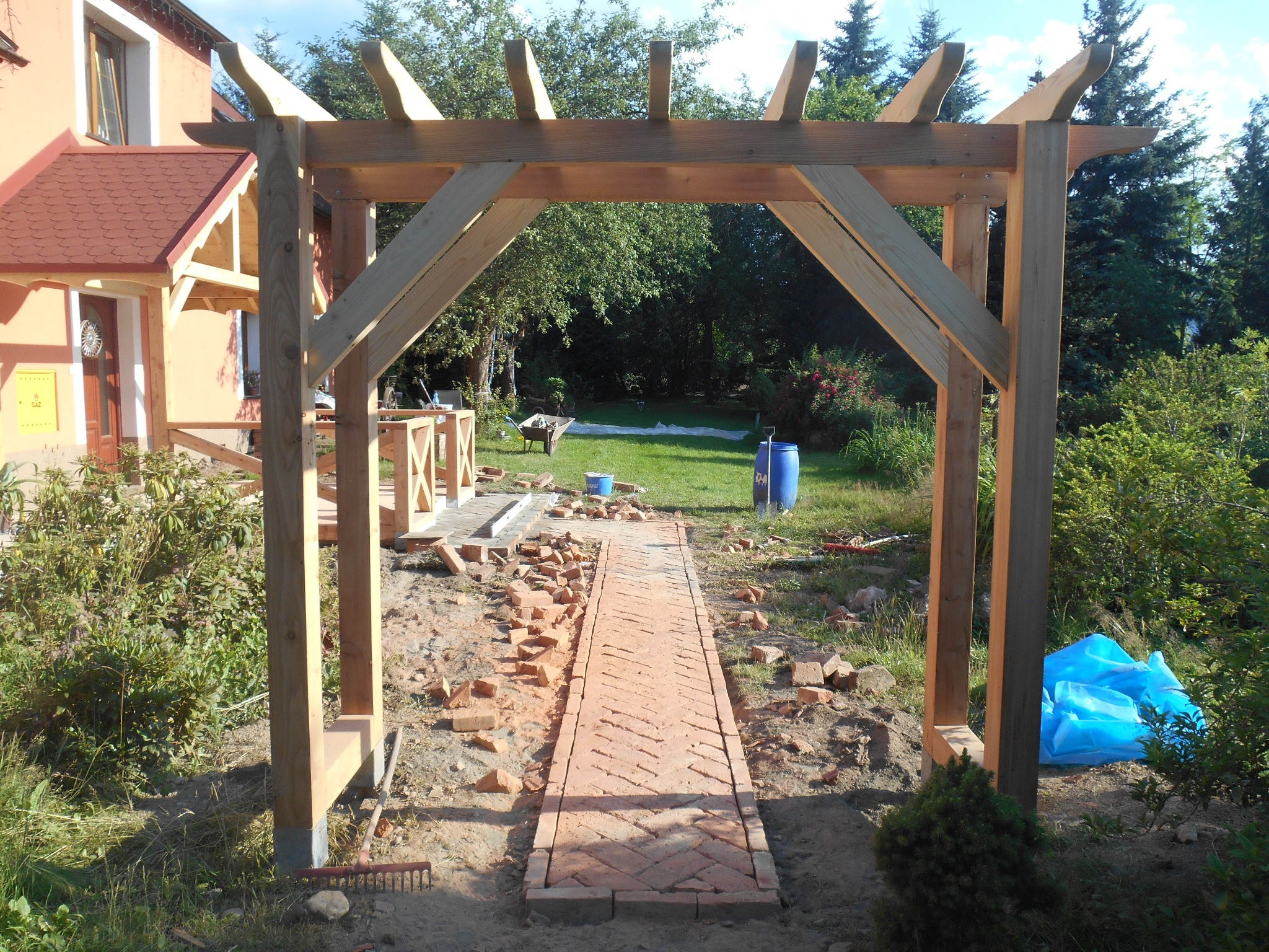 Drewniana pergola w trakcie budowy, widok na ścieżkę z cegły, elementy konstrukcyjne z surowego drewna, rozrzucone cegły, ogród w tle.