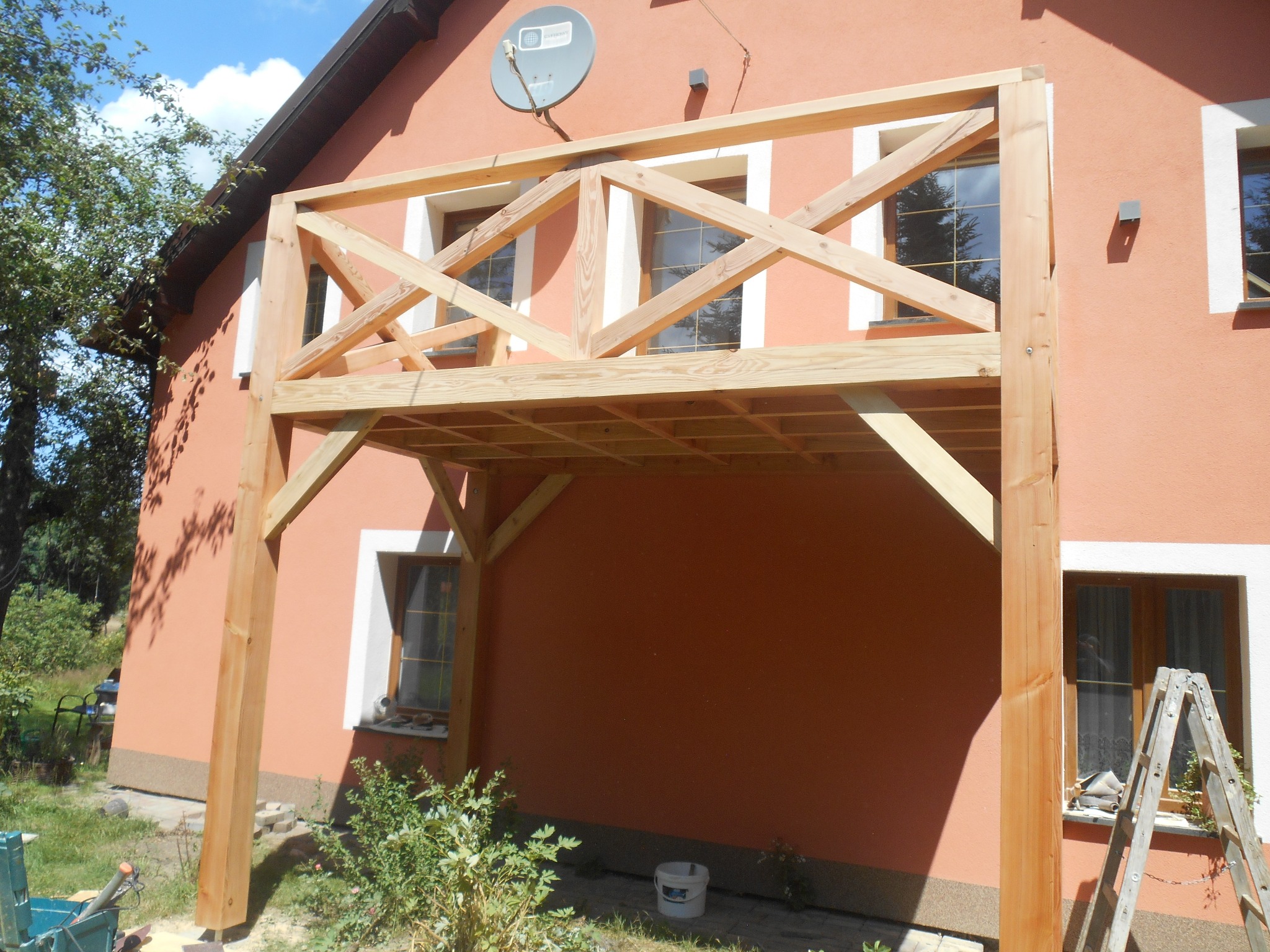 Surowy drewniany balkon z kratownicą, w trakcie budowy przy pomarańczowej elewacji domu, widoczna drabina i narzędzia.