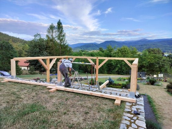 Budowa drewnianej konstrukcji tarasowej: monter w kombinezonie przy pracy, widoczne elementy konstrukcyjne i krajobraz w tle.