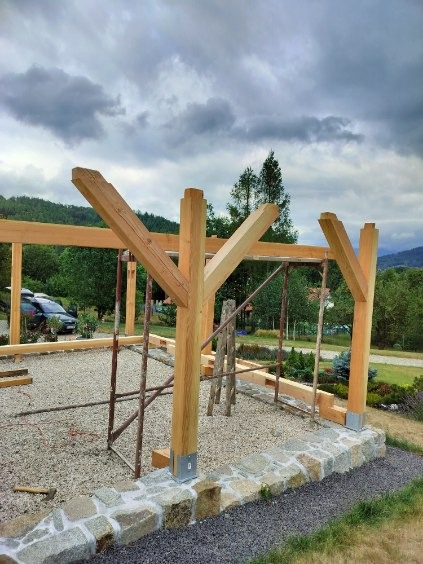 Wznoszenie konstrukcji drewnianej pergoli z widocznymi elementami montażowymi, rusztowaniami i kamiennym obrzeżem w tle krajobraz z pochmurnym niebem.