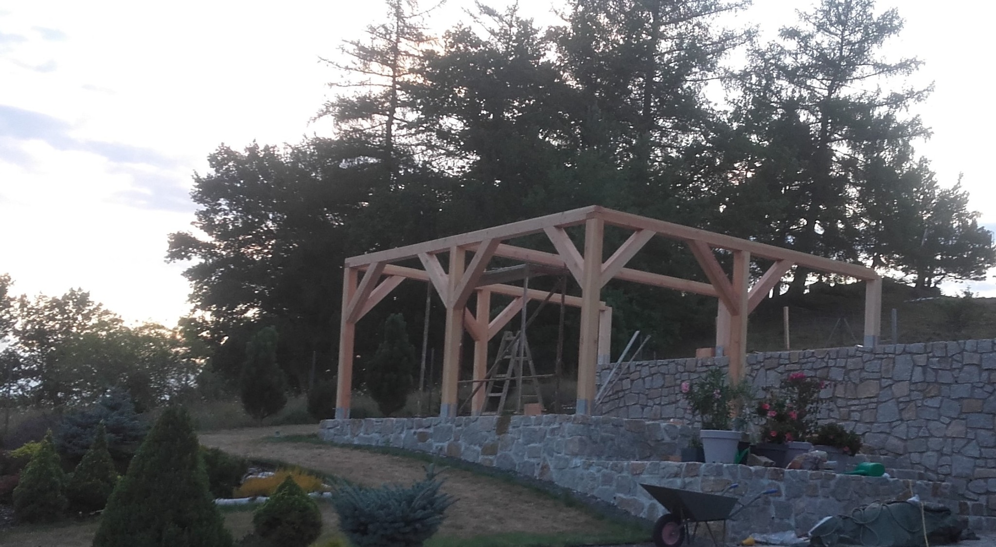 Surowa konstrukcja drewniana z belek, prawdopodobnie przyszła pergola, wznoszona na kamiennym murku oporowym z widoczną drabiną i zielenią w tle.