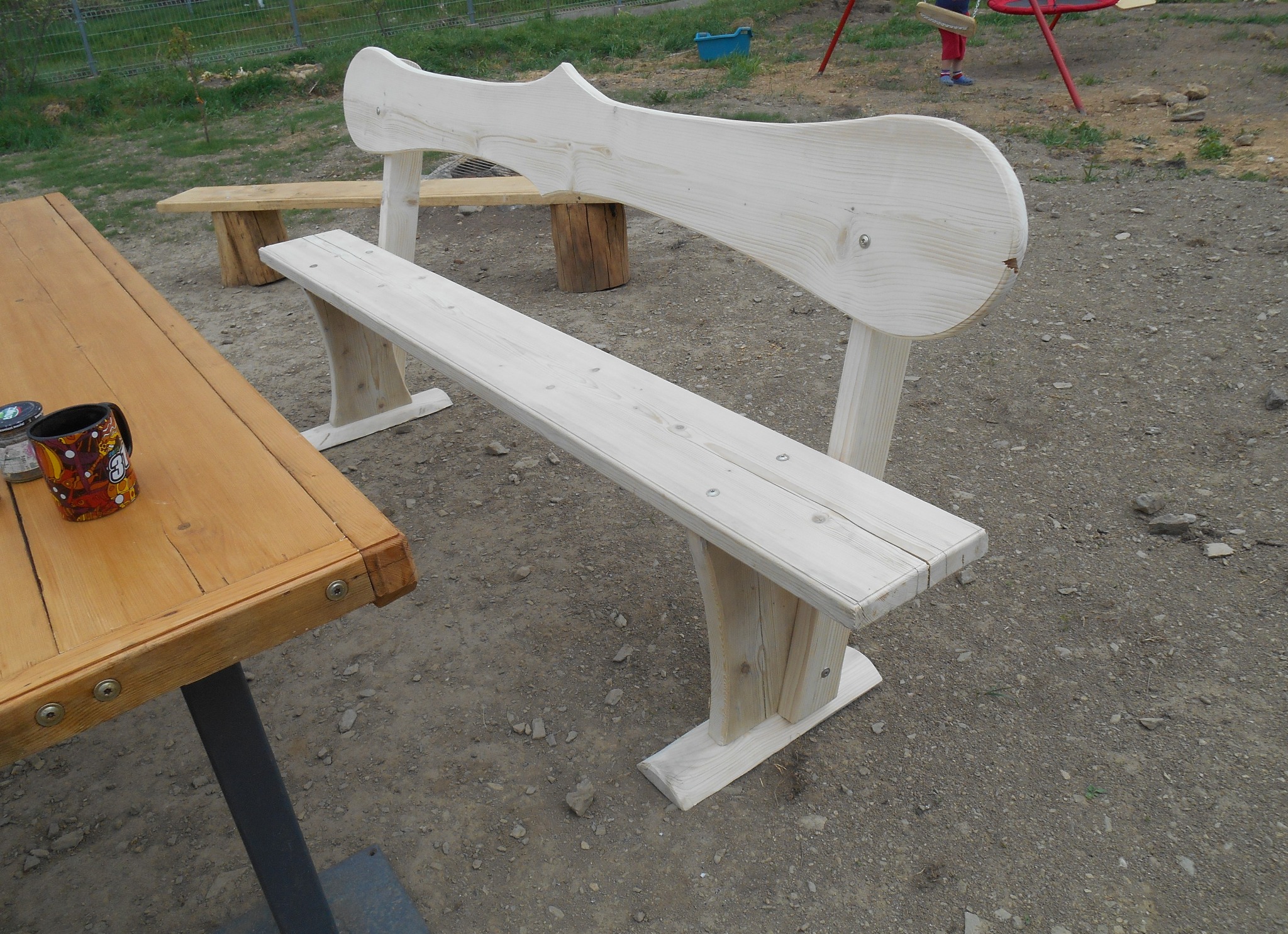 Biała, drewniana ławka z oparciem o fantazyjnym kształcie, stojąca na żwirowym podłożu, obok drewniany stół z kubkiem i mniejsza ławka w tle, na zewnątrz.