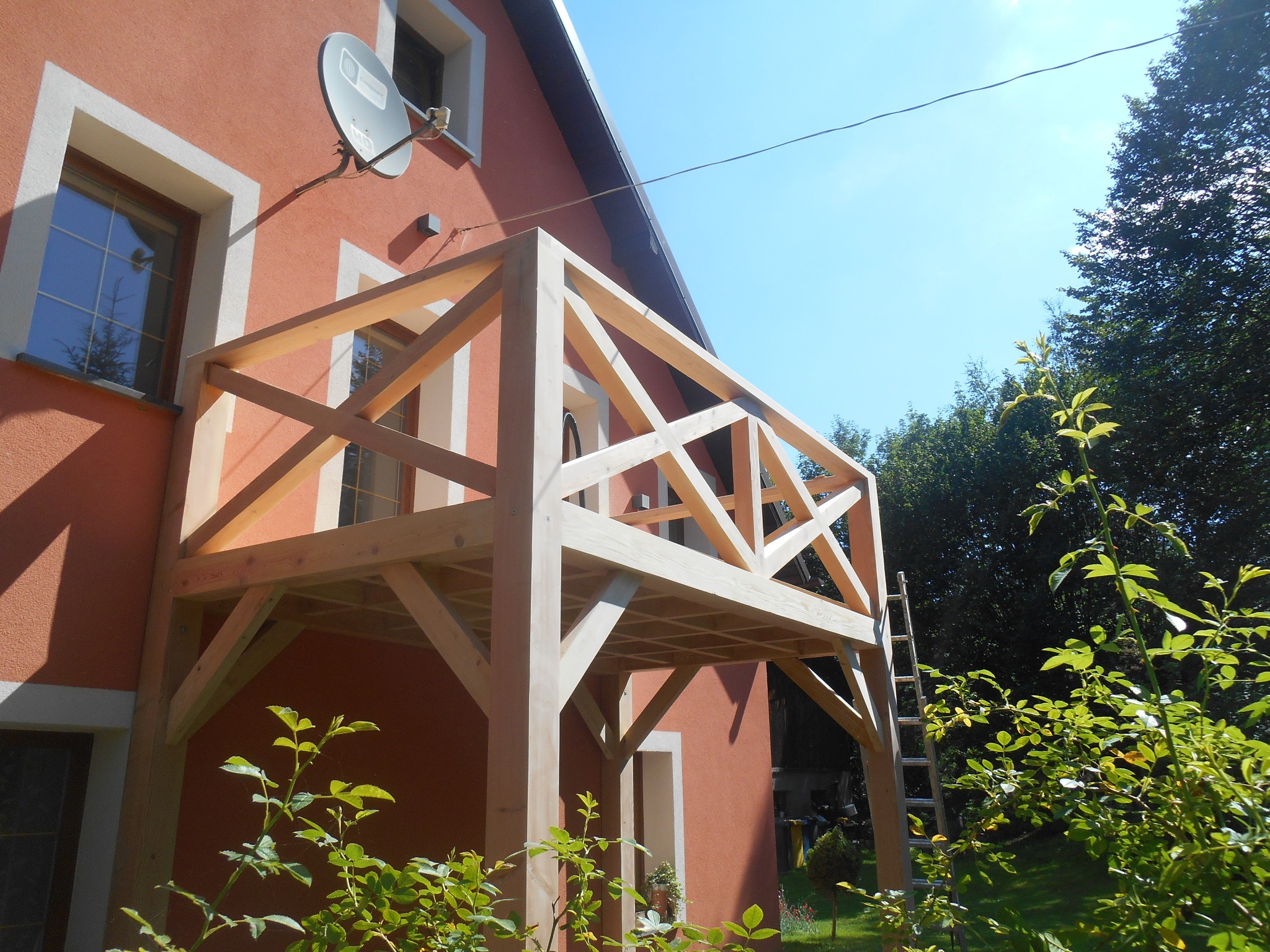 Surowy drewniany szkielet balkonu przymocowany do pomarańczowej ściany domu, widoczna drabina oparta o konstrukcję i satelita na ścianie.
