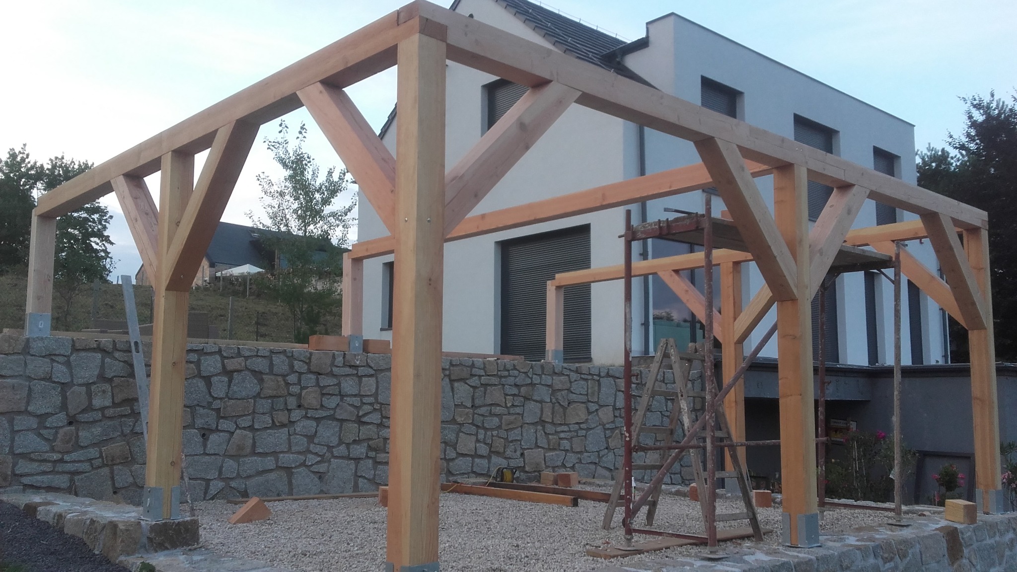 Surowa konstrukcja drewniana z elementami metalowymi, prawdopodobnie w trakcie budowy, na tle domu z szarymi roletami i kamiennego muru.