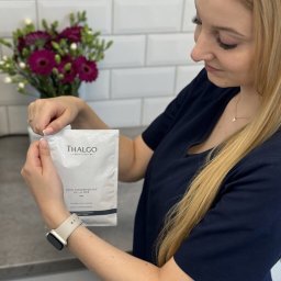 NATURAL SKIN CLINIC - salon kosmetyczny Kraków - Kobieta w uniformie otwiera saszetkę produktu Thalgo Soin Fondamental de la Mer na tle białych płytek i bukietu fioletowych kwiatów.