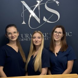 NATURAL SKIN CLINIC - salon kosmetyczny Kraków - Trzy uśmiechnięte kobiety w granatowych uniformach kosmetycznych, stoją za drewnianą ladą na tle ściany z logo salonu 'NS Natura Skin Clinic'.