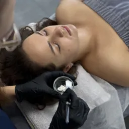 Klientka z zamkniętymi oczami leży na kozetce podczas zabiegu kosmetycznego; kosmetyczka w czarnych rękawiczkach miesza białą maskę w szklanej miseczce pędzelkiem.