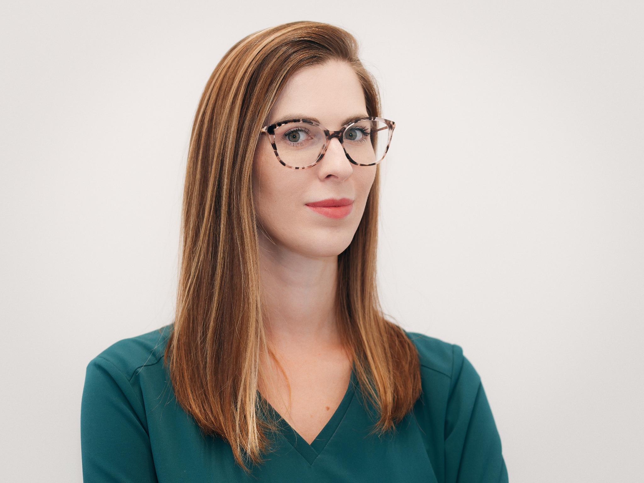 Portret kobiety w okularach o wzorzystych oprawkach, ubrana w zielony uniform, z delikatnym makijażem i brązowymi włosami na jasnym tle.