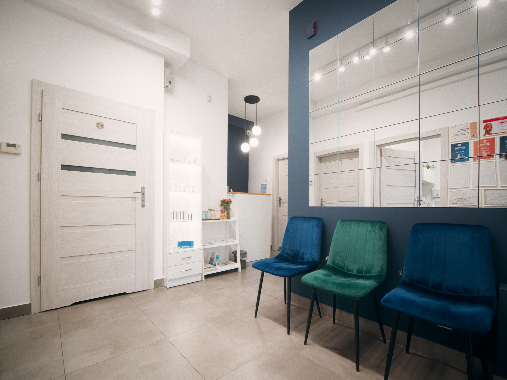 Jasne wnętrze salonu z trzema welurowymi krzesłami w odcieniach niebieskiego i zielonego, lustrzana ściana z certyfikatami i dyplomami, oraz recepcja w tle.