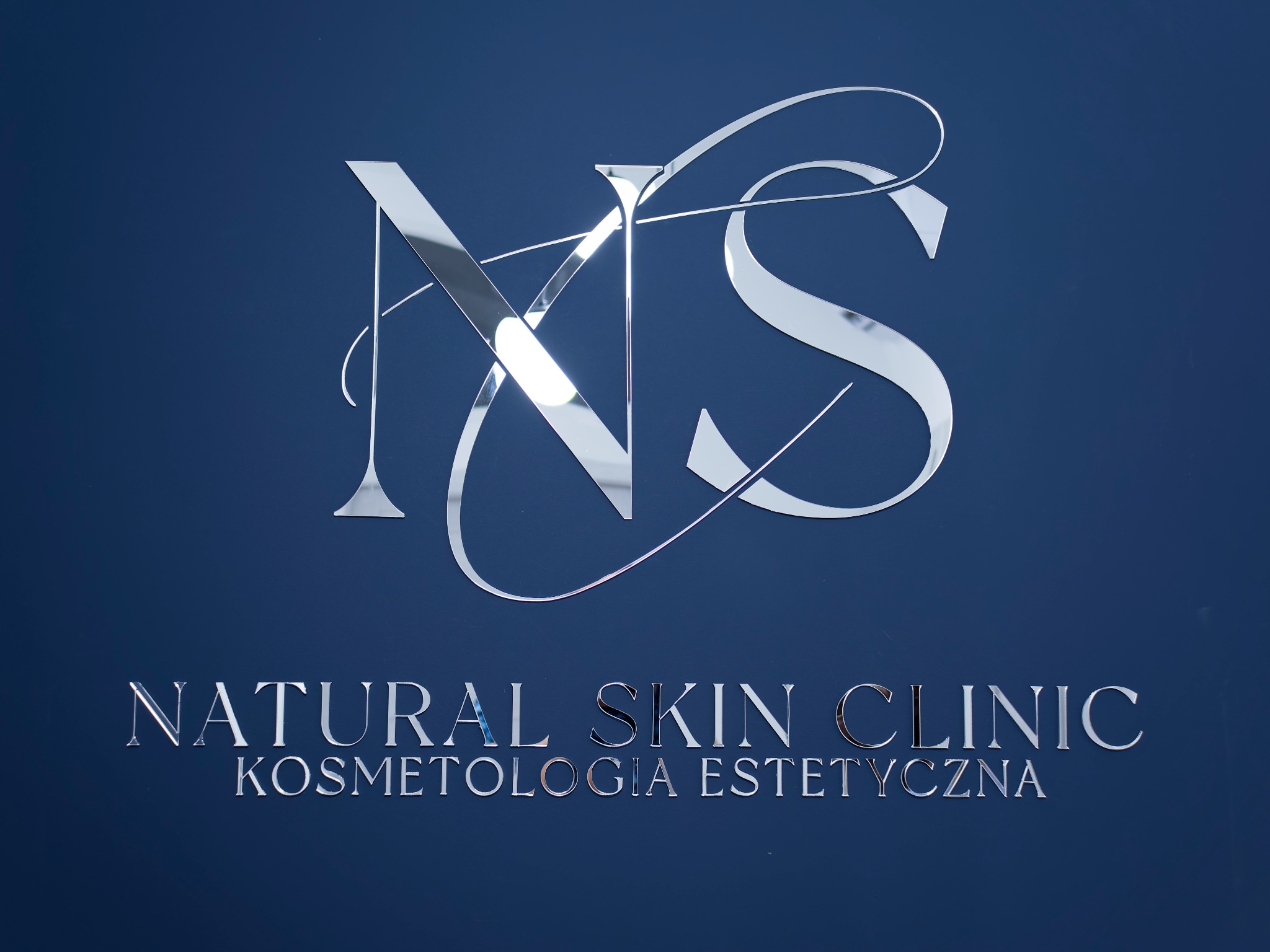 Błyszczące, srebrne logo 'NS' nad napisem 'NATURAL SKIN CLINIC KOSMETOLOGIA ESTETYCZNA' na ciemnoniebieskim tle ściany.