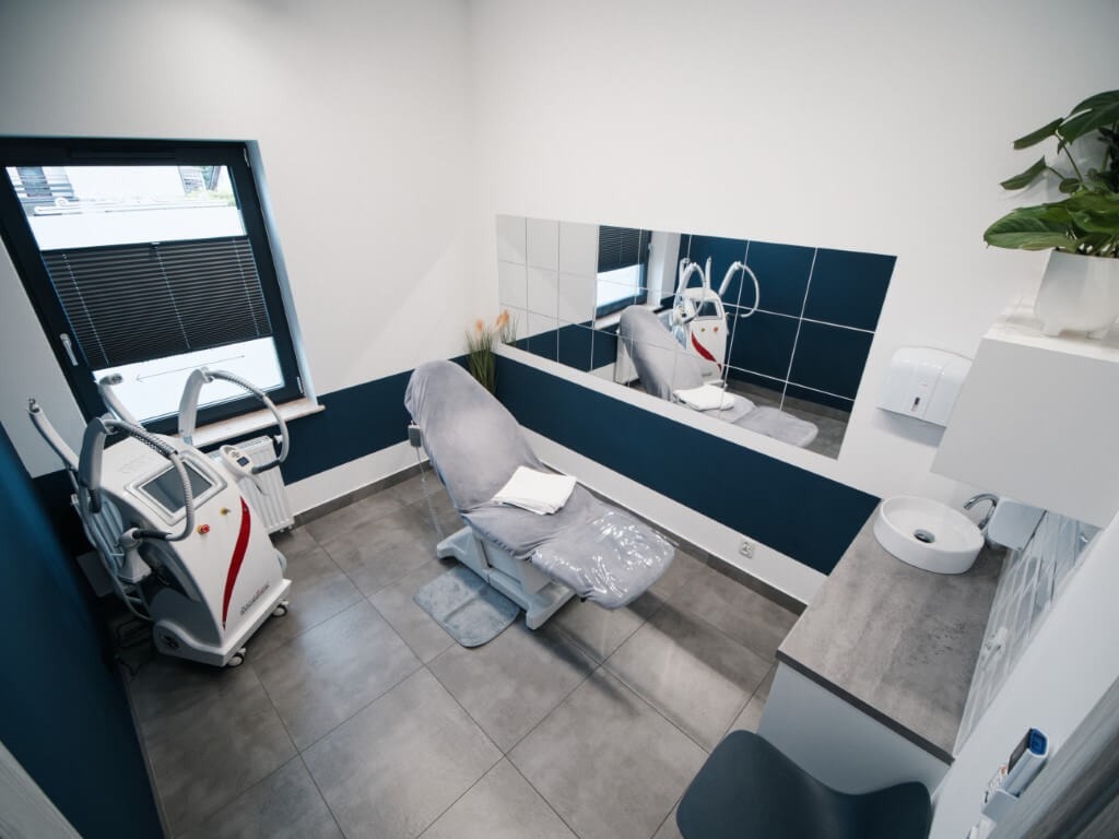 Wnętrze gabinetu z fotelem zabiegowym przykrytym szarą folią, obok urządzenie do zabiegów z wyświetlaczem i głowicą, w tle lustro i umywalka.