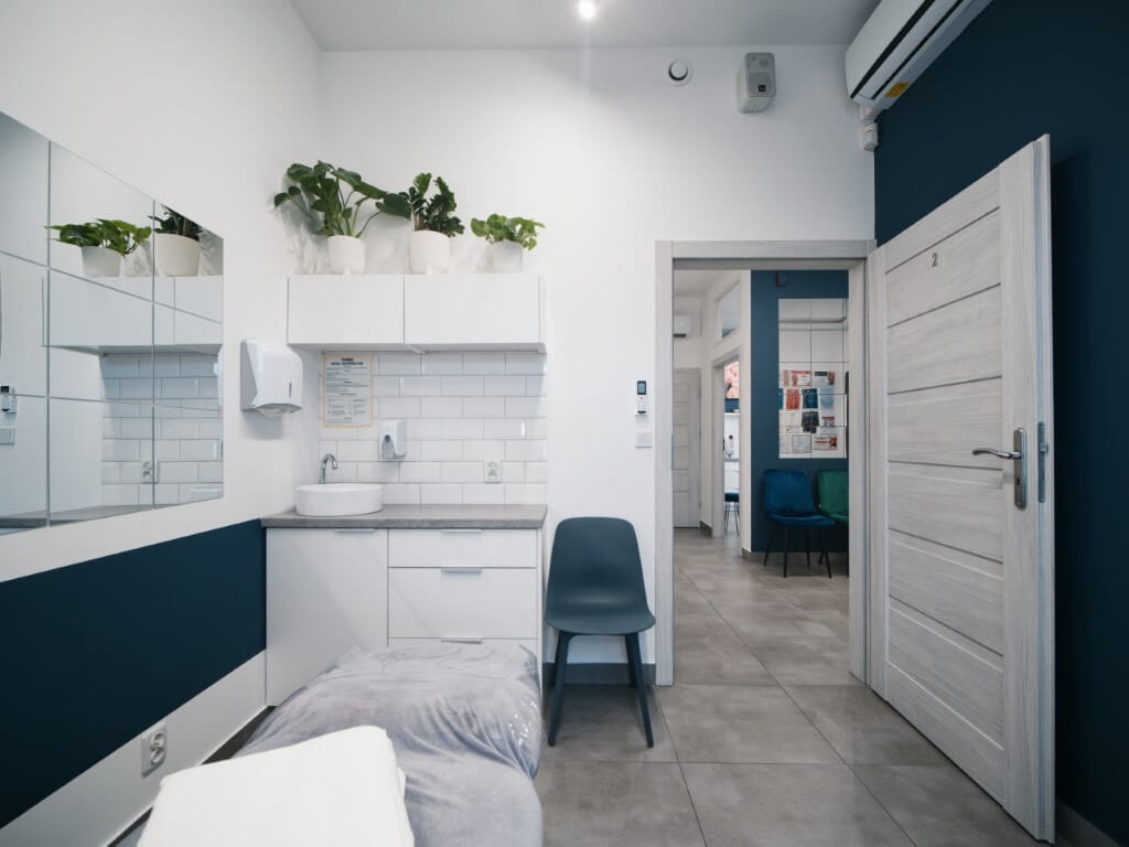 Jasne wnętrze gabinetu kosmetycznego z łóżkiem zabiegowym, umywalką, białymi szafkami, lustrem, roślinami doniczkowymi i szarym krzesłem na tle ciemnoniebieskiej ściany.