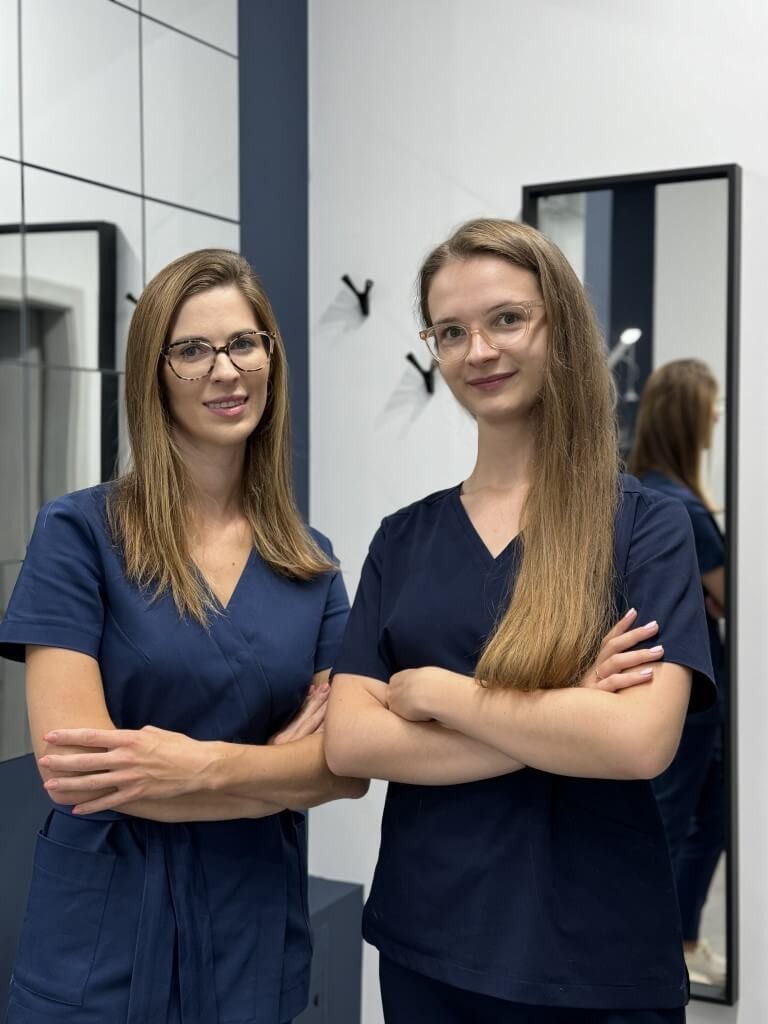 Dwie kobiety w granatowych uniformach medycznych stoją z założonymi rękami na tle jasnego gabinetu z lustrami.