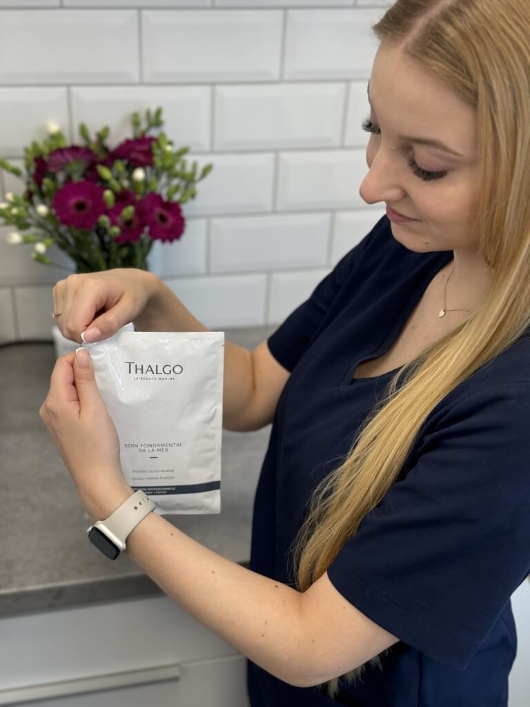 Kobieta w uniformie otwiera saszetkę produktu Thalgo Soin Fondamental de la Mer na tle białych płytek i bukietu fioletowych kwiatów.