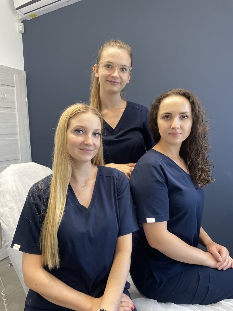 Portret trzech uśmiechniętych kobiet w granatowych uniformach kosmetycznych na tle ściany w gabinecie. Jedna z kobiet ma blond włosy, druga kręcone brązowe, a trzecia włosy spięte w kucyk i okulary.