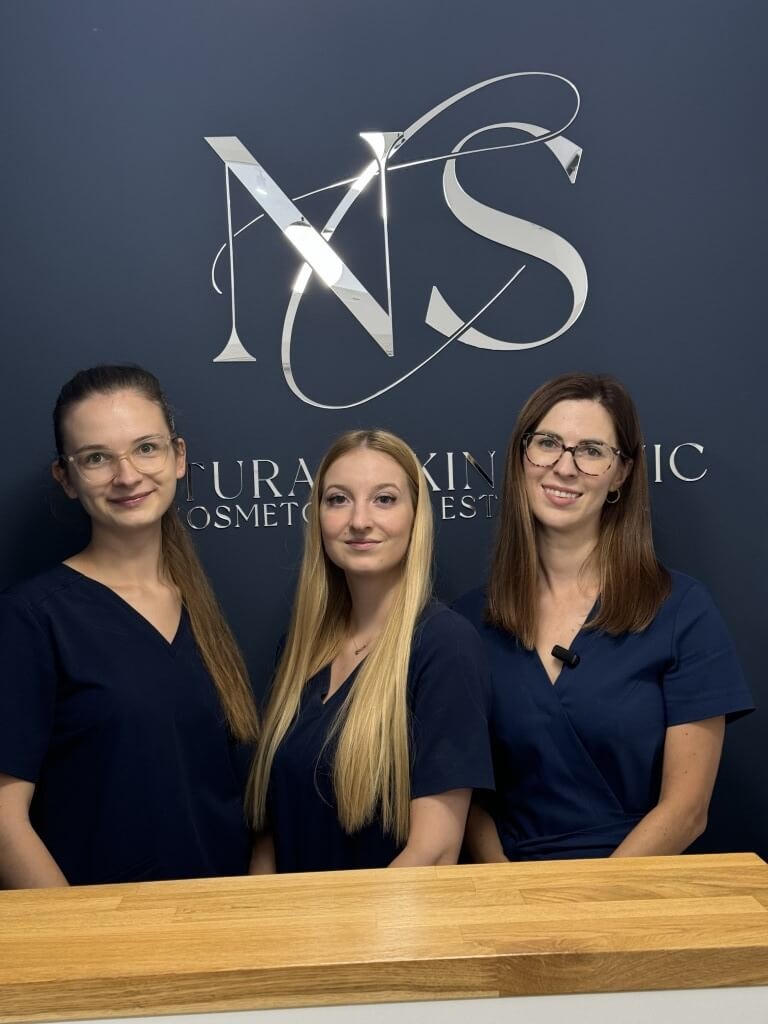 Trzy uśmiechnięte kobiety w granatowych uniformach kosmetycznych, stoją za drewnianą ladą na tle ściany z logo salonu 'NS Natura Skin Clinic'.
