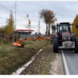 MAG-TECH Sławomir Szuhnik - Czerwony traktor z wysięgnikiem i głowicą tnącą przy rowie melioracyjnym, na tle jesiennego krajobrazu i stacji benzynowej.