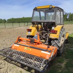 MAG-TECH Sławomir Szuhnik - Żółty traktor z pomarańczowym glebogryzem na polu, widoczne ślady pracy w ziemi i odległy las