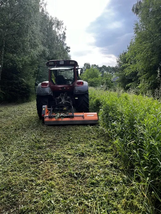 Czerwony traktor z kosiarką bijakową podczas koszenia wysokiej trawy na poboczu leśnej drogi, widok z tyłu.