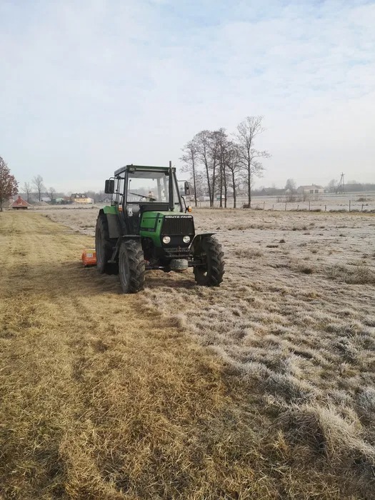 Zielony traktor Deutz-Fahr z pomarańczową kosiarką rotacyjną na zroszonym polu w chłodny poranek, widoczne budynki w oddali.