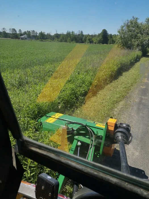 Widok z kabiny traktora na zielony teren z trzema pasami świeżo skoszonej trawy, w tle widoczne drzewa i budynek.