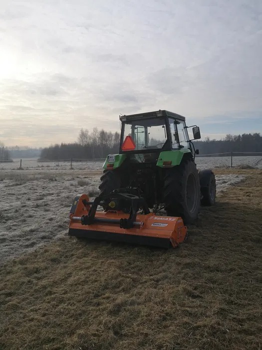 Zielony traktor z pomarańczową kosiarką bijakową marki SaMASZ na polu z niską, suchą trawą w pochmurny dzień.