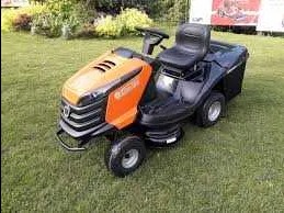 Pomarańczowo-czarny traktorek do koszenia trawy na zielonym trawniku, widoczne koła, kierownica i kosz na trawę.