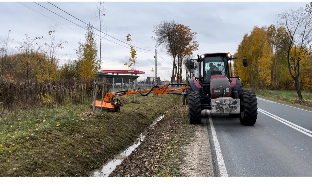 Czerwony traktor z wysięgnikiem i głowicą tnącą przy rowie melioracyjnym, na tle jesiennego krajobrazu i stacji benzynowej.
