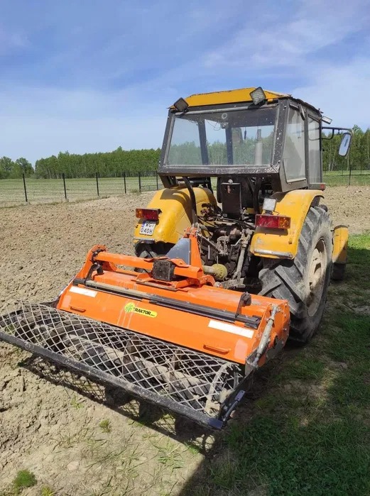 Żółty traktor z pomarańczowym glebogryzem na polu, widoczne ślady pracy w ziemi i odległy las